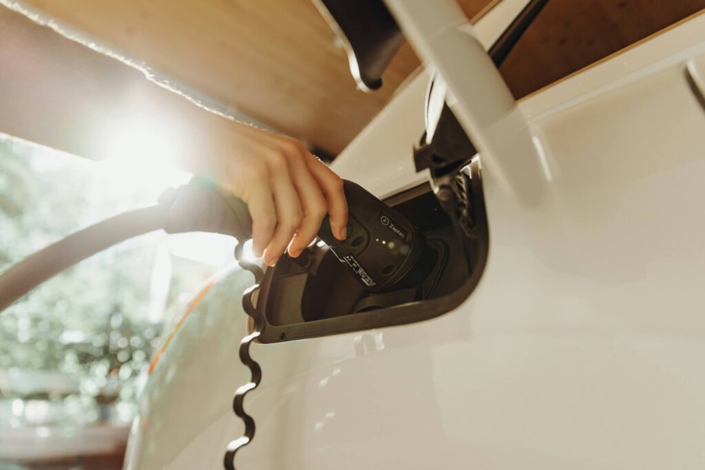Hand plugging electric car into charging port with sunlight shining in background.